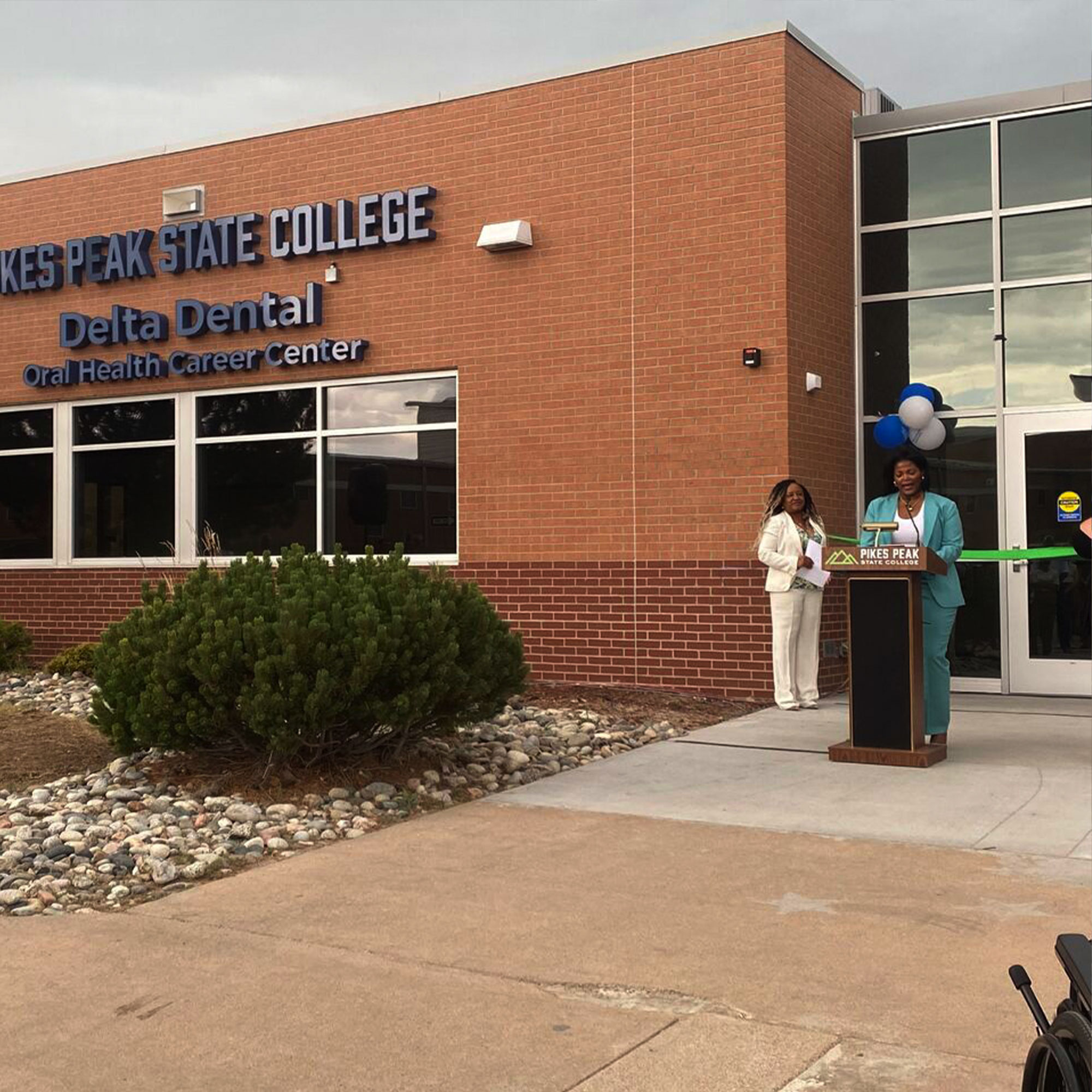 A woman speaks at a podium in front of a large building, addressing an audience with a focused expression.