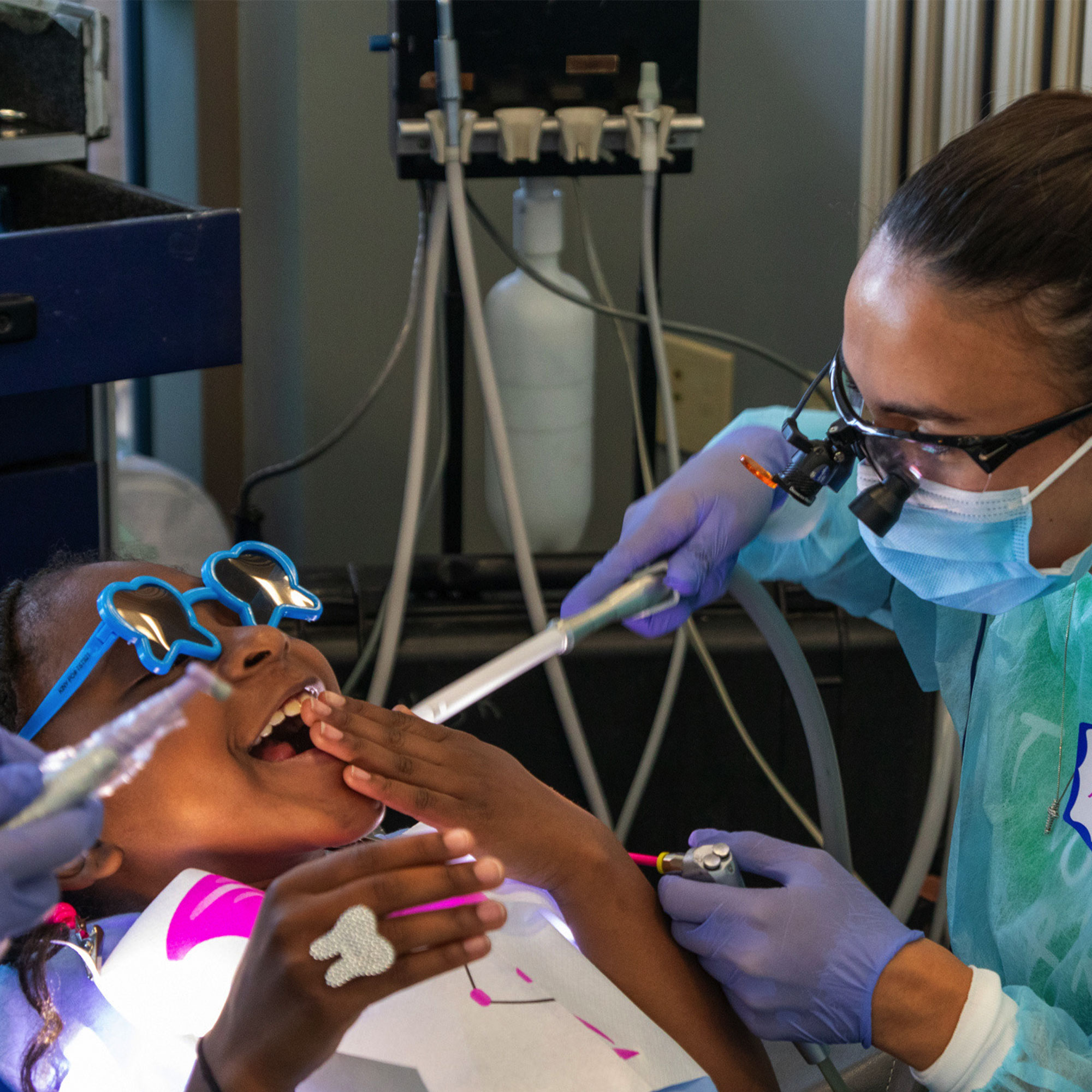 A woman in a blue coat stands beside a child receiving a dental cleaning.