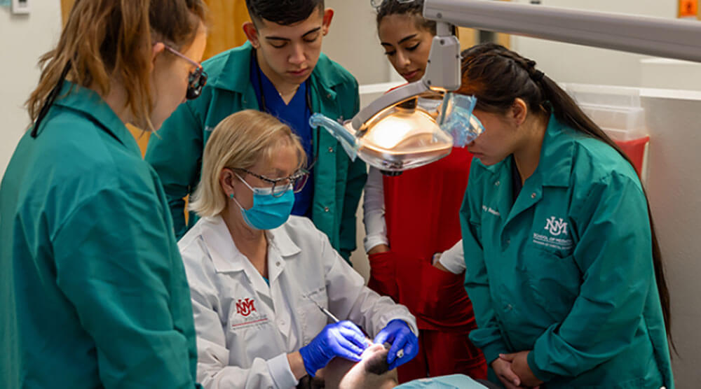 A group of students in green uniforms observes a professional dentist attending to a patient in a clinical setting.