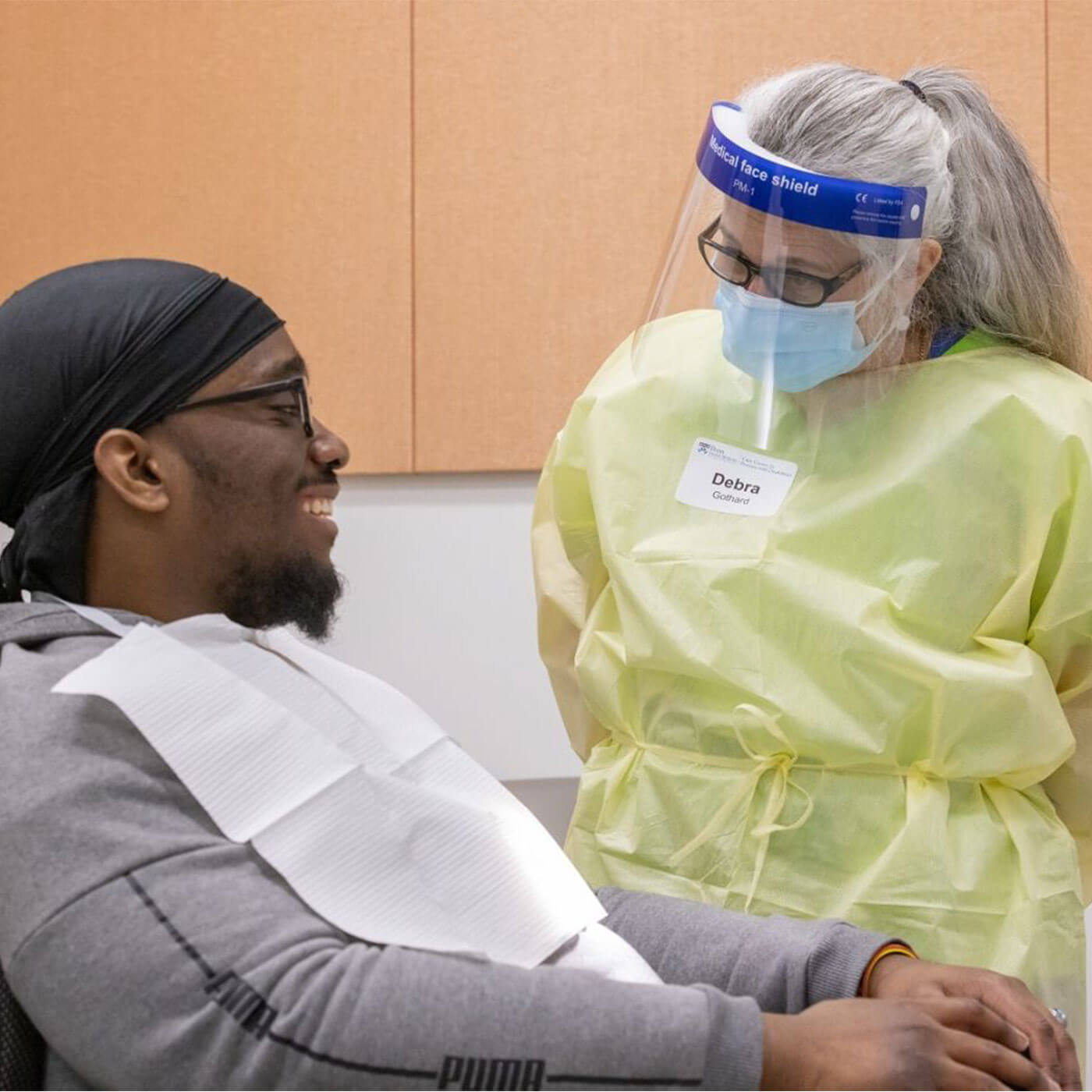 A man in a hospital gown converses with a woman wearing a face mask in a medical setting.