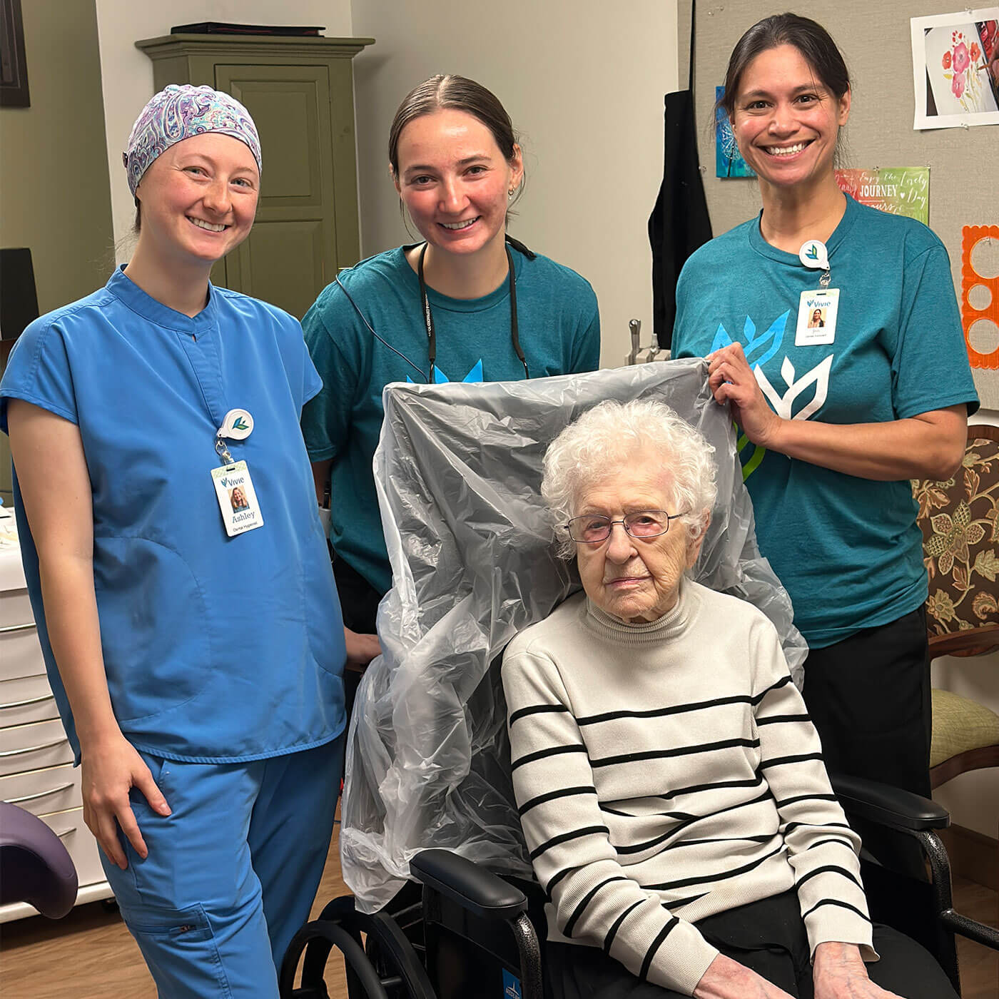 Three women in scrubs smile for a photo, with one woman seated in a wheelchair between the others.